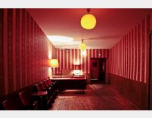 Ein schmaler, schwach beleuchteter Flur mit rot-gemusterter Tapete und Holzfußboden. Links stehen rote Polsterstühle an der Wand, daneben ein Tisch mit einer Tischlampe, während von der Decke runde Lampen hängen. Im Hintergrund führt eine geöffnete Tür in einen weiteren Raum.