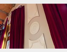 Innenansicht eines Raums mit roten, bodenlangen Vorhängen vor hohen Fenstern und dekorativen Elementen an einer cremefarbenen Wand. Eine gewölbte Decke mit freiliegenden Ziegeln ist erkennbar.