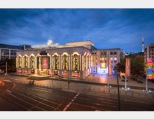 Außenansicht des Friedrichstadt-Palasts in Berlin bei Dämmerung, mit beleuchteter Fassade aus großen, bogenförmigen Fenstern und bunten Lichtelementen. Im Vordergrund eine breite Straße mit Straßenbahnschienen, im Hintergrund der Fernsehturm.