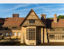 Historisches Gebäude mit Fachwerkfassade und rotem Ziegeldach, geprägt von symmetrischen Mustern und Erkern mit kleinen Fenstergittern; im Vordergrund ein gepflegter Garten mit Buchsbaumbüschen und einer niedrigen Steinmauer.