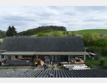 Eine ländliche Szene mit einem einstöckigen Gebäude mit grauem Ziegeldach und einer offenen Seite, auf der Baumaterialien und Gegenstände gelagert sind. Im Hintergrund befinden sich grasbewachsene Hügel mit vereinzelten Bäumen sowie eine kleine Kuhherde auf einer Weide.