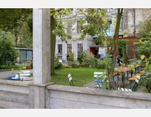 Ein grüner Außenbereich mit Holzbänken, Tischen und weißen Stühlen, umgeben von Bäumen und Pflanzen. Im Hintergrund ist ein historisches Gebäude mit hellen Fassaden und großen Fenstern zu sehen, vor dem Menschen stehen; an einigen Stellen hängen Lichterketten.