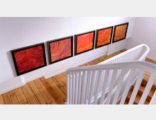 Blick in einen weißen Treppenbereich mit Holzfußboden, an dessen Wand fünf gerahmte Kunstwerke in Rot- und Orangetönen neben­einander hängen. Ein weißes Geländer führt entlang der Treppe.