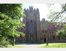 Backstein-Gotik-Kirche mit spitzbogigen Fenstern und filigranen Fassadenelementen, umgeben von grünem Laub und einem kleinen Weg im Vordergrund.