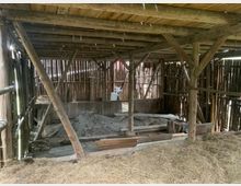 Holzstruktur einer alten Scheune mit sichtbaren Holzbalken und Zwischenräumen in den Wänden. Auf dem Boden befinden sich Heu, Bretter und ein Haufen mit Baumaterial, umgeben von einer rustikalen und unvollständig verkleideten Umgebung.