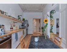 Helle Küche mit hohen Decken und Stuckverzierung, einer Arbeitsfläche aus Holz, offenen Regalen mit Gewürzgläsern und Pflanzen. Im Vordergrund ein Holztisch mit einer Sonnenblume in einer Vase, dahinter Türen, die zu anderen Räumen führen.