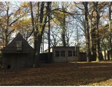 Zwei einfache Gebäude aus Beton inmitten eines herbstlichen Waldes, umgeben von hohen Bäumen mit goldbraunem Laub. Im Vordergrund bedeckt Laub den Boden, und eine schmale Laterne steht vor den Gebäuden.
