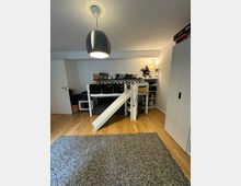 Kinderzimmer mit einem Hochbett, einer weißen Rutsche und Regalen darunter. Der Raum hat Holzboden, eine schlichte Pendelleuchte und an der Wand befinden sich Bücher und Dekorationen. Im Vordergrund liegt ein grauer Teppich.