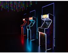 Drei retro Arcade-Spielautomaten mit leuchtenden Neonrahmen in Blau, Rot und Weiß stehen in einer dunklen Umgebung. Im Hintergrund hängen farbige, von der Decke herabhängende Dekorationsobjekte.