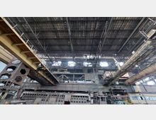 Innenansicht einer großen, verlassenen Industriehalle mit einem hohen Metallgerüstdach und mehreren Brückenkränen. Rostige Stahlkonstruktionen und verwitterte Maschinen dominieren den Raum, während Tageslicht durch Lücken im Dach einfällt.