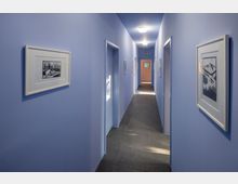 Langer, schlichter Flur mit hellblauen Wänden und dunklem Bodenbelag, dekoriert mit gerahmten Drucken an den Wänden. Türen führen zu mehreren Räumen auf beiden Seiten, und am Ende des Flurs ist eine Tür zu sehen.