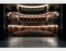 Moderner Theatersaal mit hölzernen Wänden und mehreren Sitzreihen, die sich von der Bühne bis zum hinteren Bereich erstrecken. Zwei umlaufende Balkone mit Sitzplätzen sind über den Hauptreihen angeordnet, beleuchtet von warmen, dezenten Lichtern.