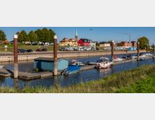 Hafenbereich mit kleinen Booten an einem Steg und einer blauen Hütte am Ufer, im Hintergrund Wohnmobile, bunte Gebäude und eine Kirche mit spitzem Turm. Die Szene ist von Grünflächen und einem klaren Himmel umgeben.