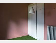 Zimmer mit rosa Wand, grünem Teppichboden und einem weißen Einbauschrank mit schwarzen Griffen. Rechts im Bild befindet sich ein Heizkörper, und eine weiße Hängelampe hängt von der Decke.