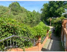 Treppenabgang mit roten Backsteinmauern und grün bewachsenen Geländern, der zu einem gepflasterten Weg entlang eines Flusses mit üppiger Vegetation führt. Im Hintergrund sind Bäume, ein klarer Himmel und ein Restaurantbereich mit Sonnenschirmen zu sehen.