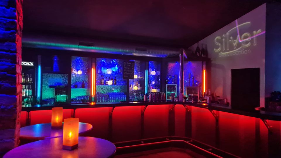 Innenraum einer modernen Bar mit U-förmigem Tresen, rot hinterleuchteter Front und beleuchteten Regalen voller Flaschen und Gläser. Neonfarbene Lichtakzente in Blau, Grün und Orange sowie kleine Tische mit Kerzenlicht prägen die Lounge-Atmosphäre.