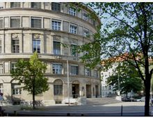 Rundes, mehrstöckiges Gebäude aus Sandstein mit symmetrischer Fensteranordnung und verzierten Fassadenelementen, umgeben von Bäumen. Im Vordergrund befindet sich ein kleiner Platz mit Treppenstufen und geparkten Fahrrädern.