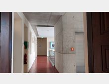 Korridor in einem modernen Gebäude mit Betonwänden, rotem Boden und einer abgehängten Decke. An der Wand befinden sich ein Feuerlöscher und eine grüne Box, während ein Bildschirm am Ende des Ganges sichtbar ist.