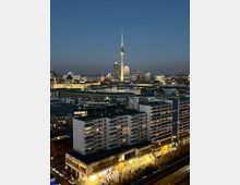 Abendansicht einer städtischen Skyline in Berlin mit dem beleuchteten Berliner Fernsehturm im Zentrum. Im Vordergrund befinden sich mehrstöckige Wohngebäude, Straßenlaternen und ein Einkaufsbereich, während der Himmel in der Dämmerung dunkelblau ist.