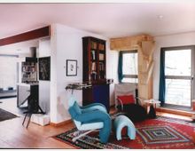 Wohnraum mit modernem Design, Holzboden und buntem Teppich. Ein ungewöhnlicher blauer Stuhl, ein Bücherregal, große Fenster mit Vorhängen und zeitgenössische Möbel bestimmen die Einrichtung, während angrenzende Räume und offene Regale im Hintergrund zu sehen sind.