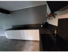 Moderne Küche mit dunklen Arbeitsplatten und weißen Schränken. An der Wand hängt ein Holzregal mit Küchenutensilien, und auf der Arbeitsfläche befinden sich eine Kaffeemaschine und Gewürzstreuer.