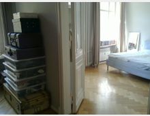 Ein helles Schlafzimmer mit Holzboden, einem Bett mit weißer Bettwäsche und einem großen Fenster mit beigefarbenen Vorhängen. Im Vordergrund links stehen stapelweise Plastikkisten und alte Koffer.