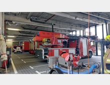 Innenansicht einer Feuerwache mit geparkten roten Feuerwehrfahrzeugen, darunter ein Drehleiterwagen. Im Vordergrund befinden sich Geräte wie eine Tragkraftspritze mit Schläuchen, und an der rechten Seite hängt Schutzkleidung für Feuerwehrleute.