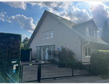 Einfamilienhaus mit Satteldach und heller Putzfassade, umgeben von Hecken und einem Metallzaun. Im Vordergrund ein gepflasterter Eingangsbereich mit einem Basketballkorb und bepflanzten Blumenbeeten.