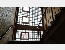 Treppenhaus in einem Mehrfamilienhaus mit rau verputzten Wänden und Metallgeländern, aufgenommen von unten nach oben. Große Glasbausteinfenster und einige rechteckige Klappfenster lassen viel Tageslicht in den Innenraum.