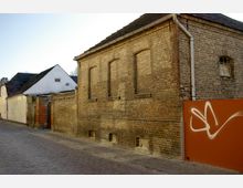 Straßenszene mit Kopfsteinpflaster, flankiert von einem gelblichen Ziegelgebäude mit zugemauerten Fenstern und einem weißen Haus im Hintergrund. Rechts ist eine orangene Wand mit einem Graffiti sichtbar.