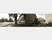 Straßenszene in einem kleinen Dorf mit alten Gebäuden; links eine asphaltierte Straße mit vereinzelten Autos und Bäumen, rechts einfache, teils verputzte Häuser mit Ziegel- und Steinmauern sowie ein Hof, der von einer Mauer eingefasst ist.