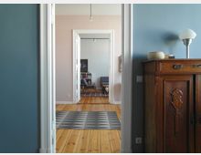 Ein Blick durch einen Flur in eine helle Wohnung mit Holzboden, pastellfarbenen Wänden und weißen Türrahmen. Im Hintergrund befindet sich ein Wohnzimmer mit einem Bücherregal, zwei Sesseln und einem gemusterten Teppich.