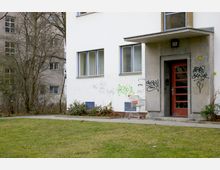 Mehrfamilienhaus mit weißer Fassade und blauen Fensterrahmen, teilweise mit Graffiti bemalt; ein rotes Eingangstor mit Vordach und ein leerer Einkaufswagen stehen im Vordergrund. Im Hintergrund ist ein weiterer Wohnblock sowie kahle Bäume sichtbar.