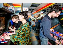 Innenansicht einer Retro-Spielhalle mit mehreren Arcade-Automaten. Menschen spielen an den Automaten, umgeben von bunten Lichtern und Spiegeln an der Wand, während der Raum mit einem karierten Boden gestaltet ist.