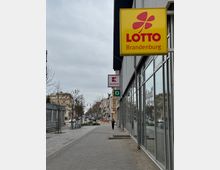 Straßenszene mit breitem Gehweg, gesäumt von Schaufenstern eines modernen Gebäudes rechts und vereinzelten kleinen Bäumen links. Im Vordergrund ein gelbes „Lotto Brandenburg“-Schild, dahinter weitere Hinweise auf Geschäfte, darunter ein „Kaufland“-Logo. Im Hintergrund sind Wohn- und Geschäftshäuser sowie eine befahrene Straße sichtbar.