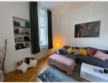Wohnzimmer mit grau bezogenem Ecksofa, dekoriert mit bunten Kissen und einer rosa Decke. Im Hintergrund ein großes Fenster mit dunkelgrünen Vorhängen, ein beleuchteter Stehlampen-Schirm sowie ein Bücherregal, das in die weiße Wand integriert ist. Der Boden besteht aus Parkett, und ein gemusteter Teppich liegt unter einem weißen Couchtisch.