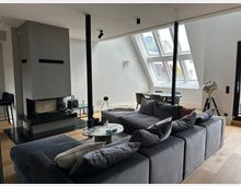 Modernes Dachgeschoss-Wohnzimmer mit großer grauer Sofalandschaft, rundem Couchtisch und freistehendem Kamin auf schwarzer Steinplatte. Schräge Decke mit großen Dachfenstern und Blick nach draußen, dahinter ein Esstischbereich sowie ein Stativ-Scheinwerfer neben einer Glastür.