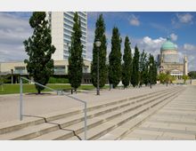 Moderne Außenanlage mit breiten Treppen, einer Reihe von säulenartigen Bäumen und Gehwegen. Im Hintergrund ein Bürogebäude sowie eine Kirche mit einer grünen Kuppel und klassischer Architektur.