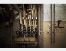 Ein Kellerraum mit alten Heizungsrohren, mehreren verrosteten Ventilen und Druckmessgeräten; die Wände weisen abblätternden Putz und sichtbare Alterungsspuren auf.