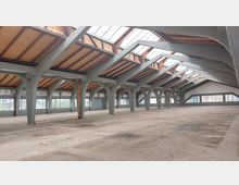 Leerer Innenraum einer großen Industriehalle mit freiliegendem Dachgebälk aus Holz und Stahlträgern. Oberlichter aus Glas und Fensterreihen entlang der Wände sorgen für natürliches Licht, während der Betonboden staubig und ungenutzt ist.