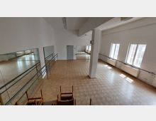 Ballett- oder Tanzstudio mit hellem Holzboden, großen Wandspiegeln und Ballettstangen entlang der Wände. Drei große Fenster auf der rechten Seite lassen Tageslicht in den Raum, der von einer zentralen Säule geteilt wird.