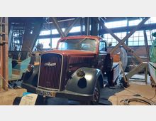 Historischer Lastwagen des Modells „Opel Blitz“ in einer industriellen Halle mit großen Fenstern und Metallträgern. Im Vordergrund steht eine Infotafel, die den LKW beschreibt.