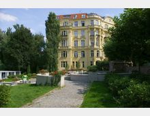 Gelbes, mehrstöckiges Wohngebäude im Stil des Historismus mit dekorativen Fassadenelementen, umgeben von einem kleinen Park mit gepflegten Grünflächen, Blumenbeeten und gepflasterten Wegen. Im Vordergrund sind Sträucher, eine Frau und Blumenkübel mit roten Blüten zu sehen.