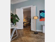 Heller Innenraum mit Holzboden und hohen weißen Doppeltüren, die in einen angrenzenden Raum mit Regalen und Lampenlicht führen. Im Vordergrund sind ein kleiner Tisch mit Kosmetikprodukten, eine Zimmerpflanze und eine Stehlampe zu sehen, an der Wand ein abstraktes Gemälde.