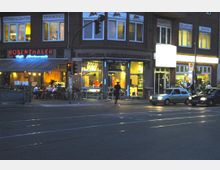 Straßenszene bei Dämmerung mit einem Gebäude, das verschiedene Geschäfte beherbergt, darunter ein Café-Restaurant „Rosenthaler“ mit Außenbestuhlung und ein Imbiss „Grill Hähnchen“ mit beleuchteter Auslage. Im Vordergrund sind eine Ampel, geparkte Autos und Straßenbahnschienen sichtbar.