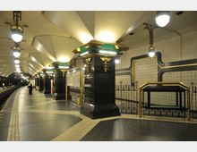 U-Bahn-Station mit Art-déco-Elementen, gekennzeichnet durch große säulenartige Strukturen mit schwarz-goldenem Design, beleuchtet von hängenden Lampen. Der geflieste Boden und die Wände enthalten geometrische Muster mit goldenen Akzenten, während ein Bahnsteig mit Schienen und Sitzbereichen zu sehen ist.