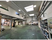 Industriehalle mit grünem Betonboden, hohen Decken und großem Oberlicht. Auf der linken Seite befinden sich Metallregale und Palettenlager, auf der rechten Seite Regale mit Farbdosen sowie freiliegende Lüftungsrohre an der Wand.