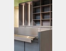 Minimalistischer Innenraum mit grauen Einbaumöbeln, die aus Schubladen und offenen Regalfächern bestehen. Im Vordergrund eine glatte Theke mit einer ausklappbaren Metallablage; die Rückwand zeigt vertikale Lichtakzente.