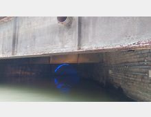 Blick auf einen gemauerten Kanal unter einer rostigen Metallbrücke, mit Wasser an der Unterseite und einem leuchtenden blauen Licht am Ende des Tunnels.