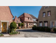 Straßenszene in einem Wohngebiet mit Einfamilienhäusern, die Putzfassaden und rote Ziegeldächer haben. Im Vordergrund ein gepflasterter Gehweg, Hecken und ein Hauseingang mit gelber Tür und kleinem Vordach.